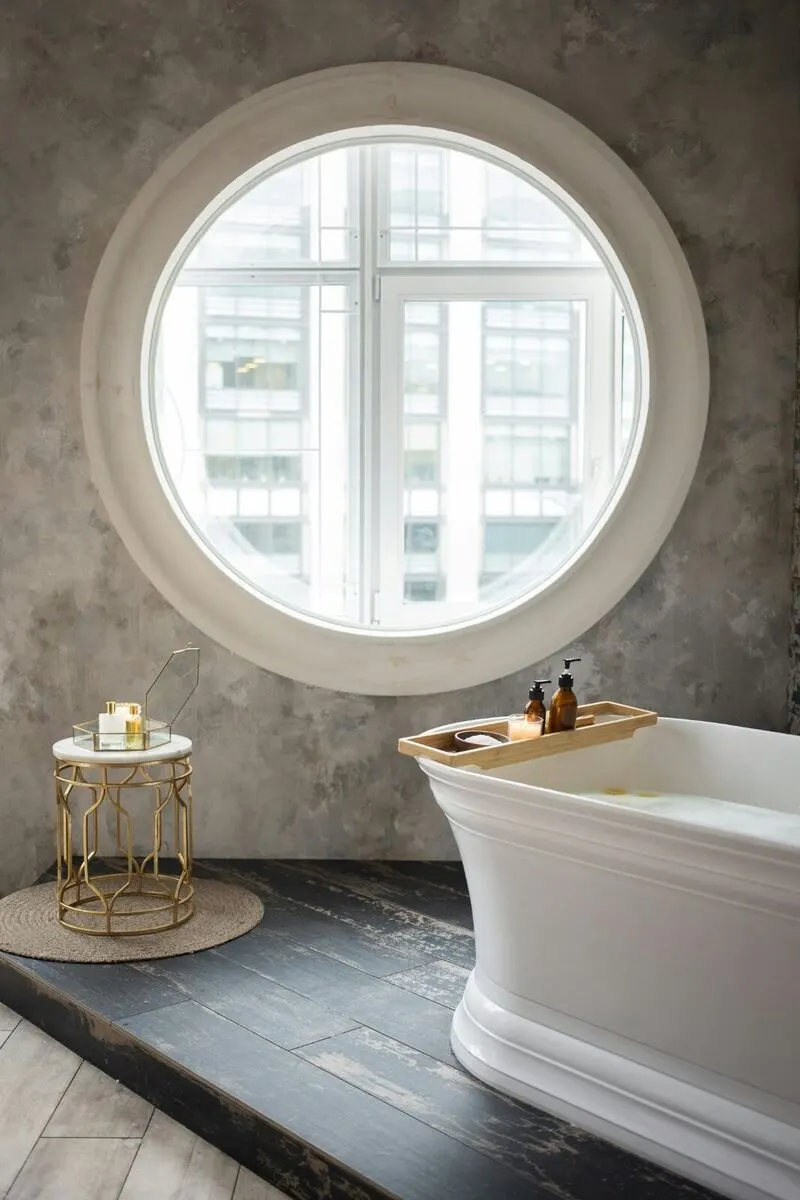 Freestanding tub with porthole window concrete walls and brass accents